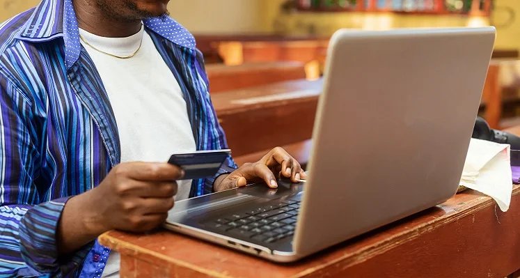 An African man looking at his laptop while holding his bank card.