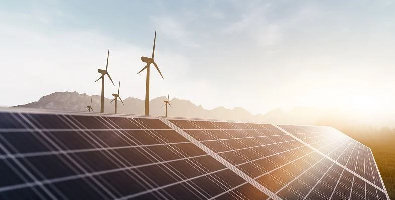 Solar panels with the sun shining on them with wind turbines in the background.