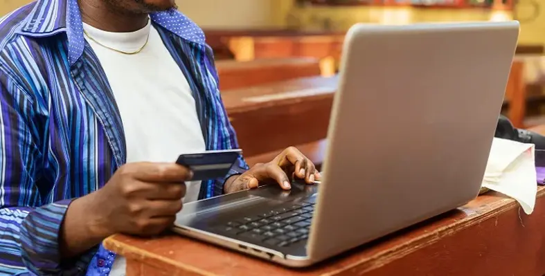 An African man looking at his laptop while holding his bank card.