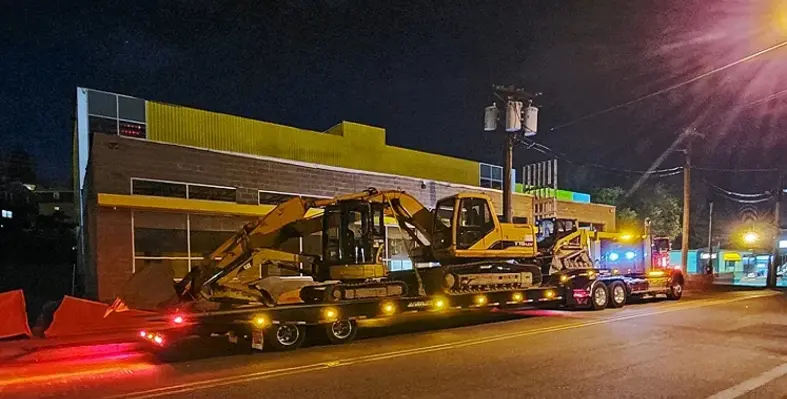 A travelling axle unit carrying construction equipment through a street.