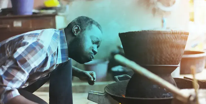 A_man_blowing_on_a_fire_to_help_him_cook_food.